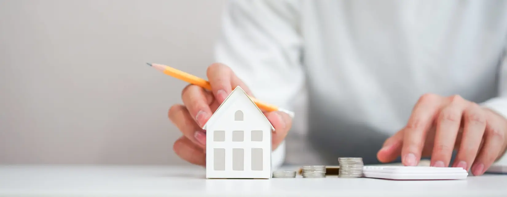 Person mit Miniatur Haus aus Papier neben Münzen und einen Taschenrechner