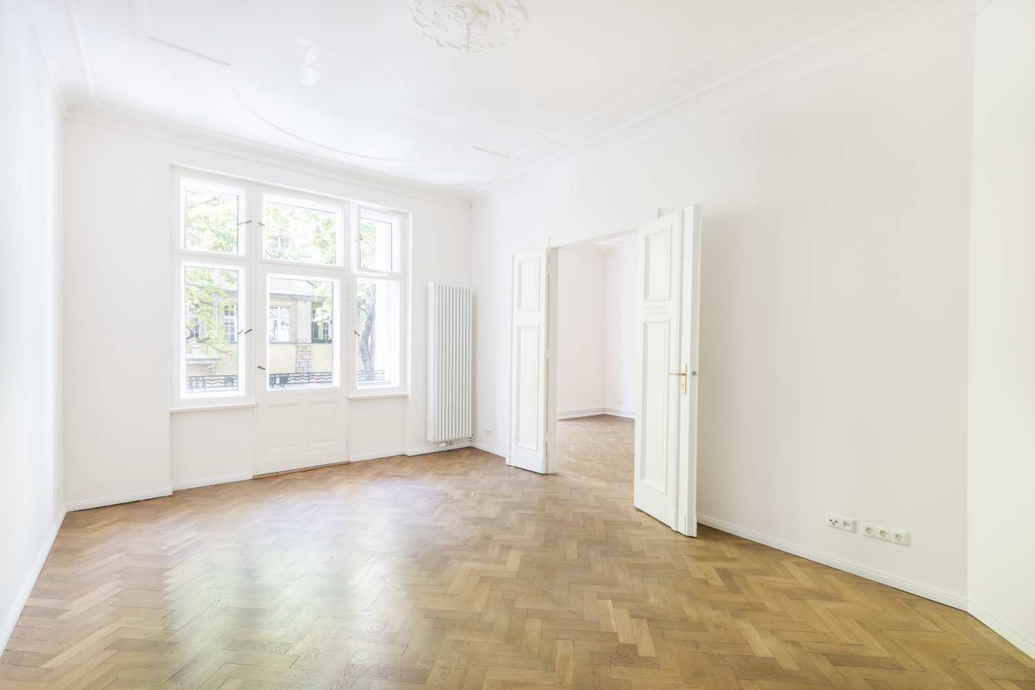 Geräumiges Wohnzimmer in einem Altbau mit hohen Decken, doppelten Flügeltüren und Fischgrätparkett, das eine helle und elegante Atmosphäre schafft.