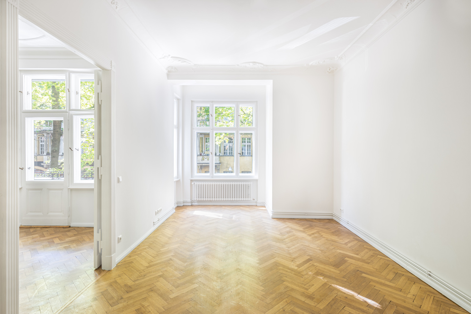 Renovated old building room with large windows, herringbone parquet flooring and ornate ceiling moldings, combining historical charm with modern living comfort.