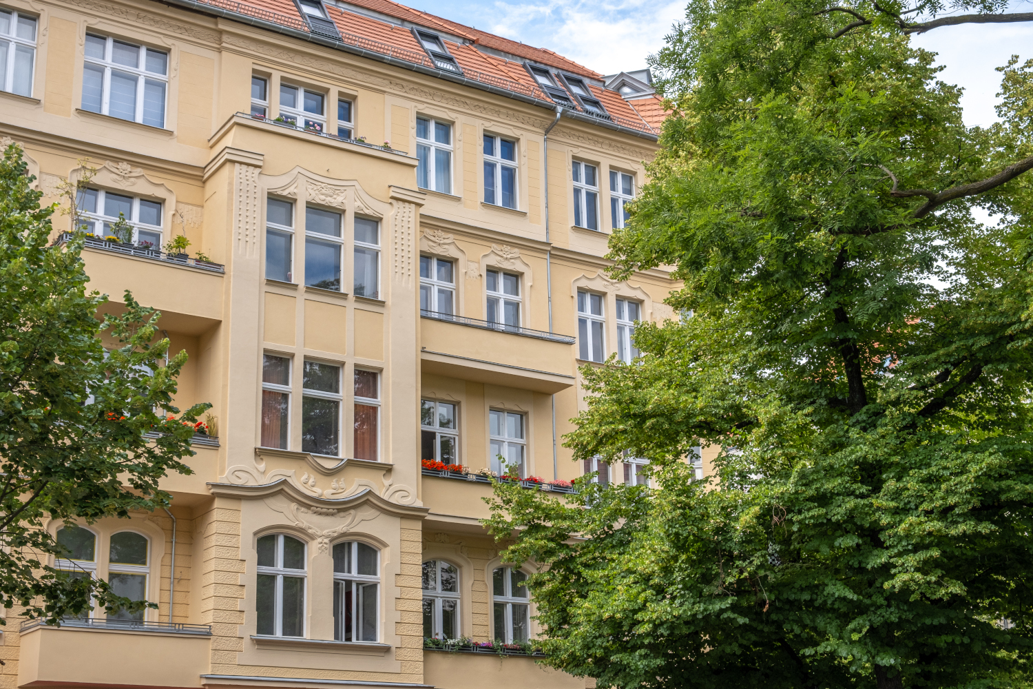 Altbaufassade eines sanierten Mehrfamilienhauses mit aufwendigen Verzierungen und klassischen architektonischen Details, umgeben von grünen Bäumen.