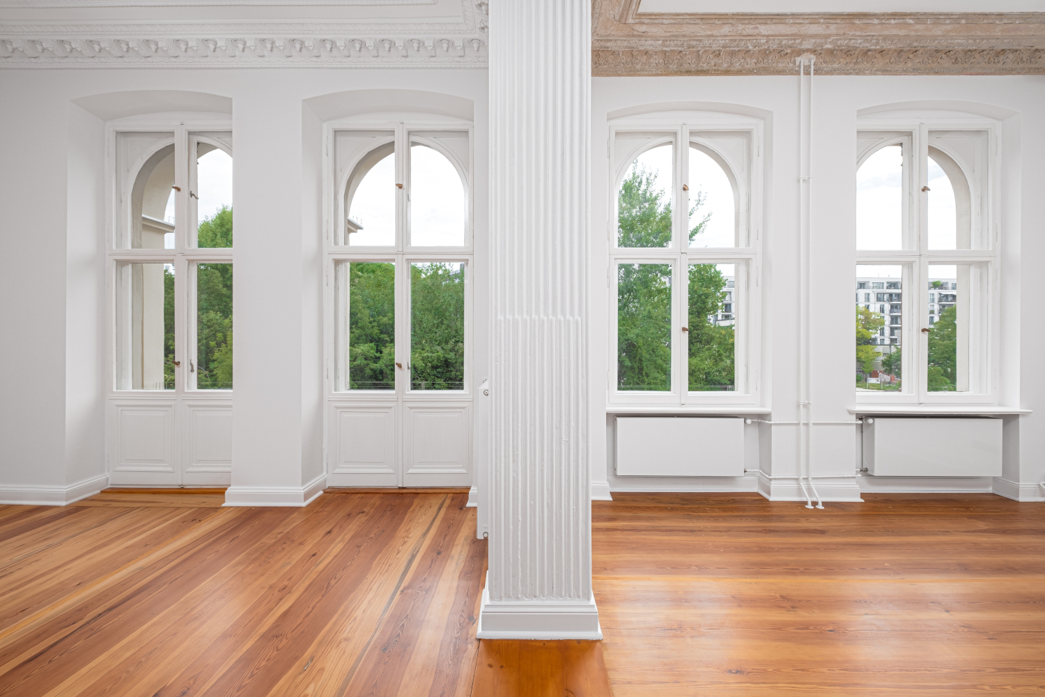 Bright old building room with original stucco decorations on the ceiling and large windows offering a view of the surrounding green landscape.”